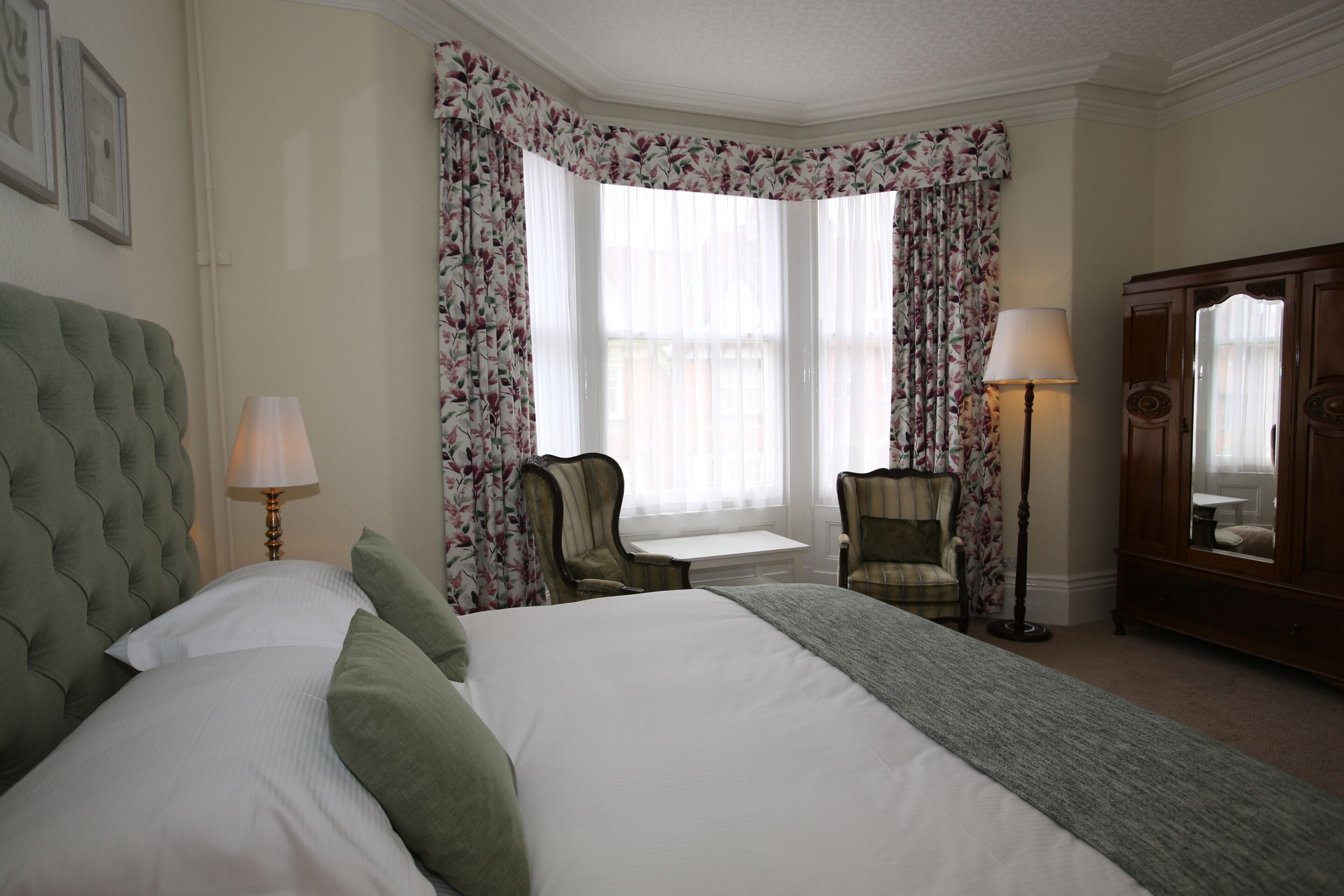 A hotel room with a large bed, a desk, a chair, a lamp, and a wardrobe.