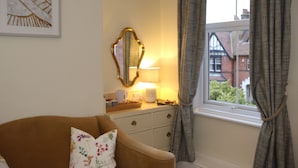 A room with a brown sofa, a mirror, a lamp, a window with curtains, and a framed picture on the wall. - The Cordelia (Scarborough)