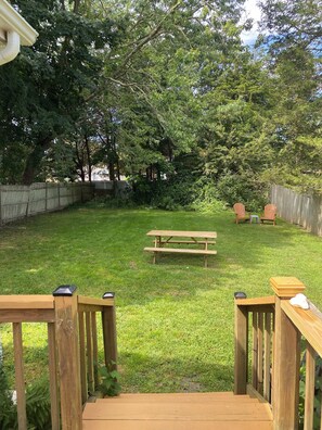 Enjoy a serene outdoor space with a wooden deck, picnic table, and Adirondack chairs surrounded by lush greenery.