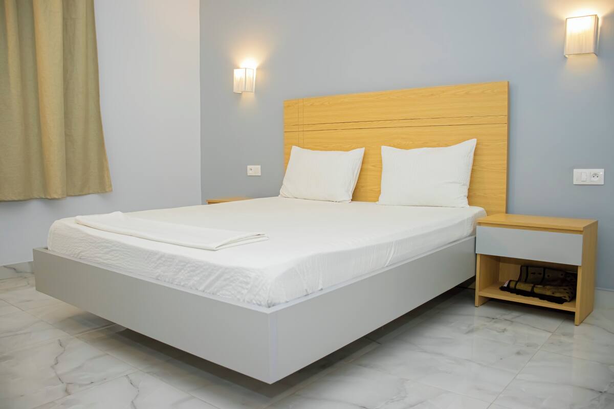 A modern hotel room with a large bed, a wooden headboard, white bedding, two bedside tables, and a nightstand with a lamp.