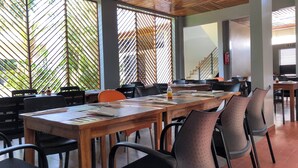 Un vaste espace dédié à la salle à manger, avec de longues tables en bois et des chaises modernes, de grandes fenêtres équipées de stores verticaux, et un escalier en arrière-plan.