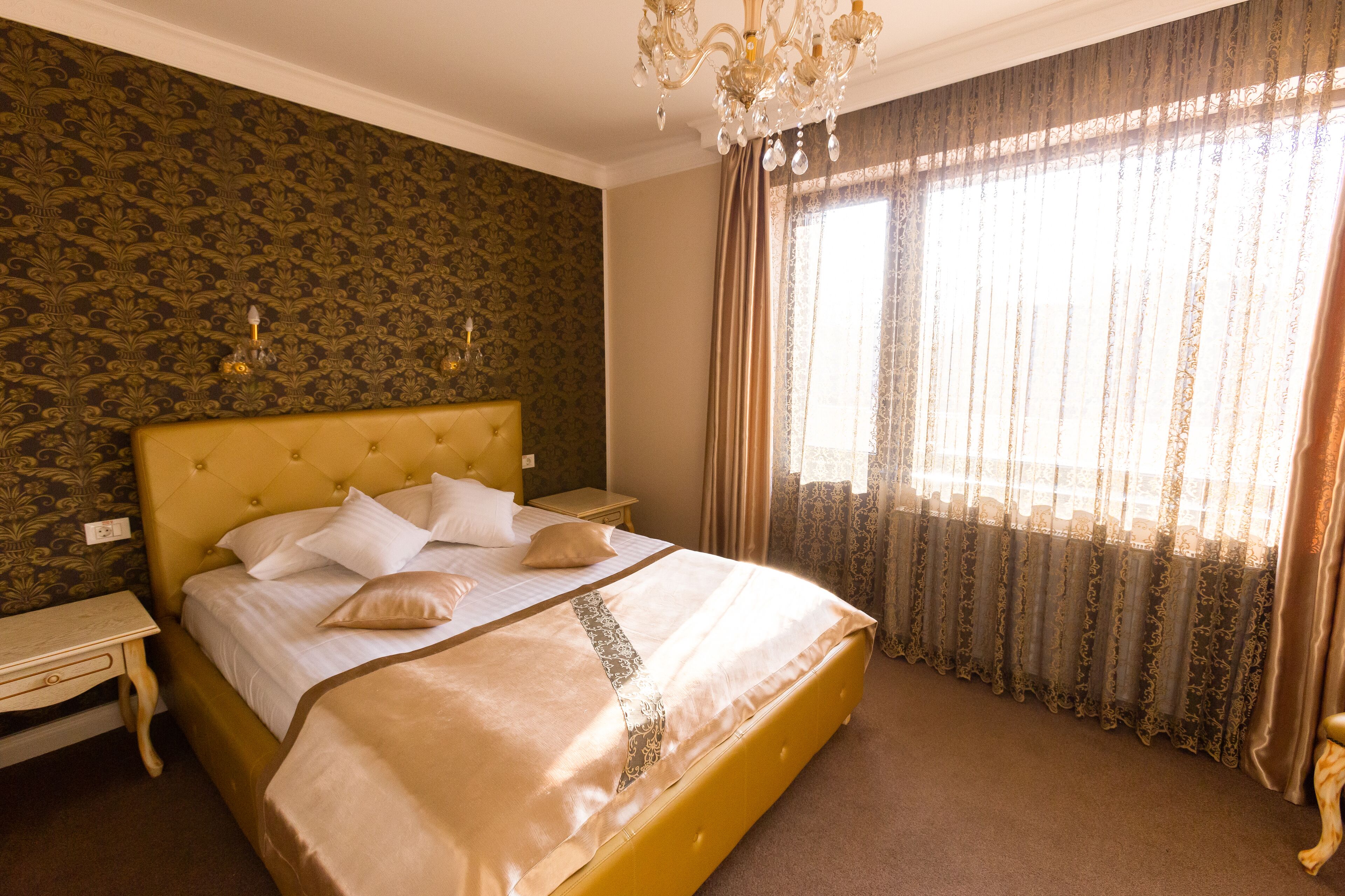 A hotel room with a large bed, a nightstand, a chandelier, and a window with curtains.