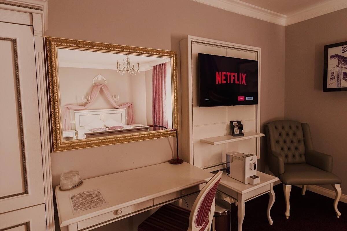 A hotel room with a vanity area, a television displaying the Netflix logo, a chair, and a small table with a telephone and a box.