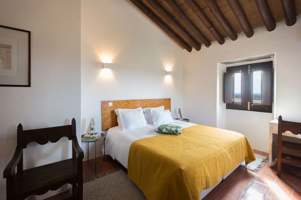 A bedroom with a wooden headboard, a bed with white bedding and a mustard-colored blanket, two bedside lamps, a wooden chair, a small table, and a window with a view.