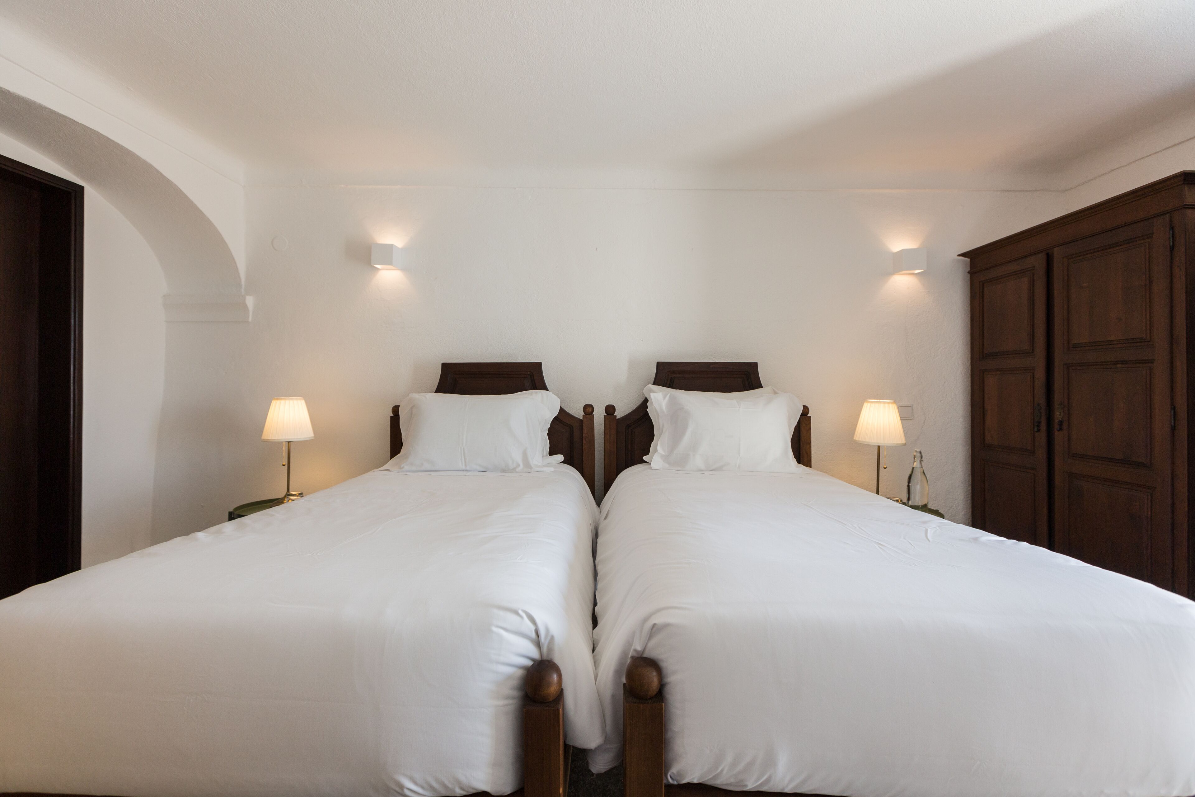 A double bed with white linens and wooden headboards, two bedside lamps, and a wooden wardrobe.
