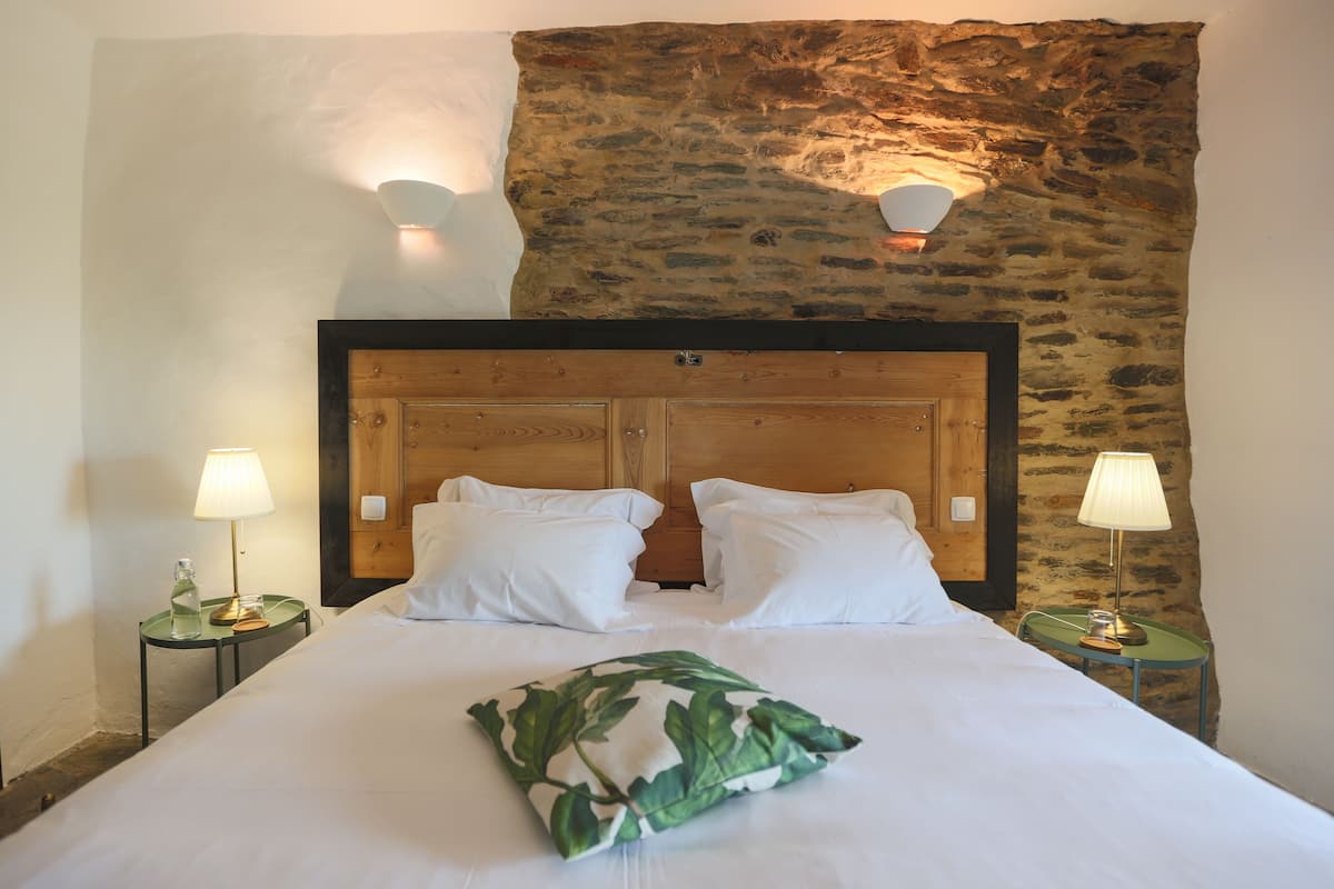 A neatly made bed with white bedding and pillows, flanked by two bedside tables with lamps, set against a rustic stone wall.