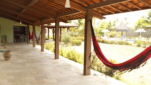 Un patio cubierto con una hamaca, suelo de piedra y vistas a una piscina y jardín.