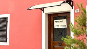 Un edificio con facciata rossa, una porta in legno con un cartello che dice 'Affittasi' e una piccola finestra con persiane.