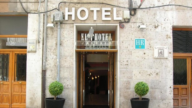 The entrance to El Hotel Jacobeo, featuring a stone facade, two potted trees, and a sign indicating the hotel's name. - Hotel Jacobeo (Burgos)