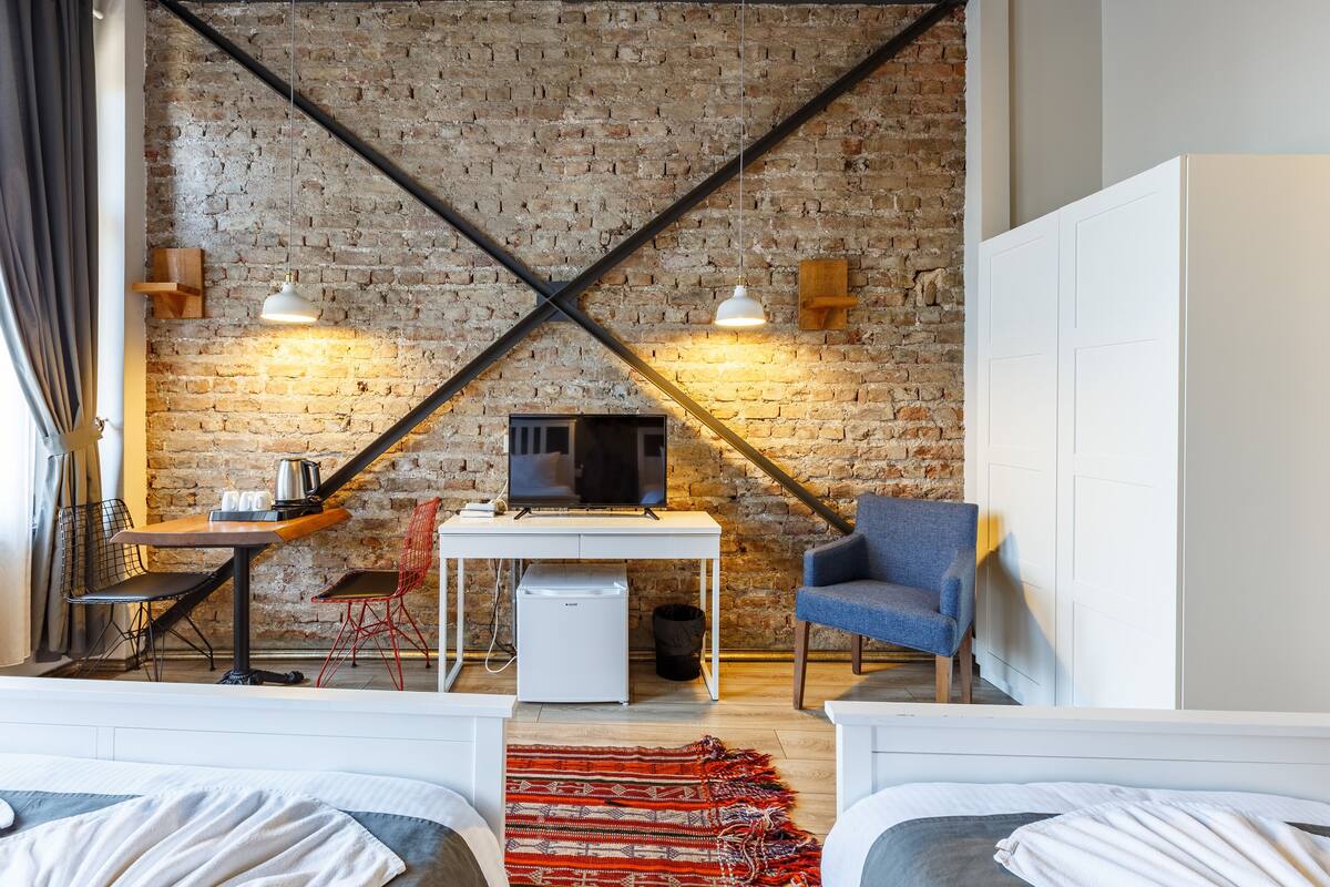 Una habitación con pared de ladrillo, un escritorio blanco con televisor, una silla azul y dos camas.