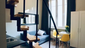 A modern kitchen with a built-in sink, a wooden staircase, a grey sofa with a polka dot pillow, a yellow chair, and a white cabinet. - GOLDA apartments (Lodz)