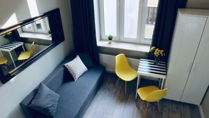 A modern room with a grey sofa, a small desk with a chair, a radiator, and a window with curtains. - GOLDA apartments (Lodz)