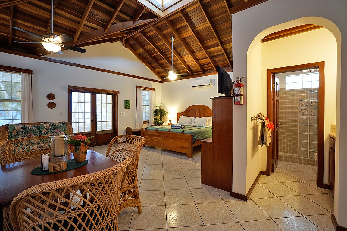 Une pièce spacieuse avec un plafond en bois, un lit doté d’une tête de lit en bois, une table à manger avec des chaises en osier et un petit coin cuisine.