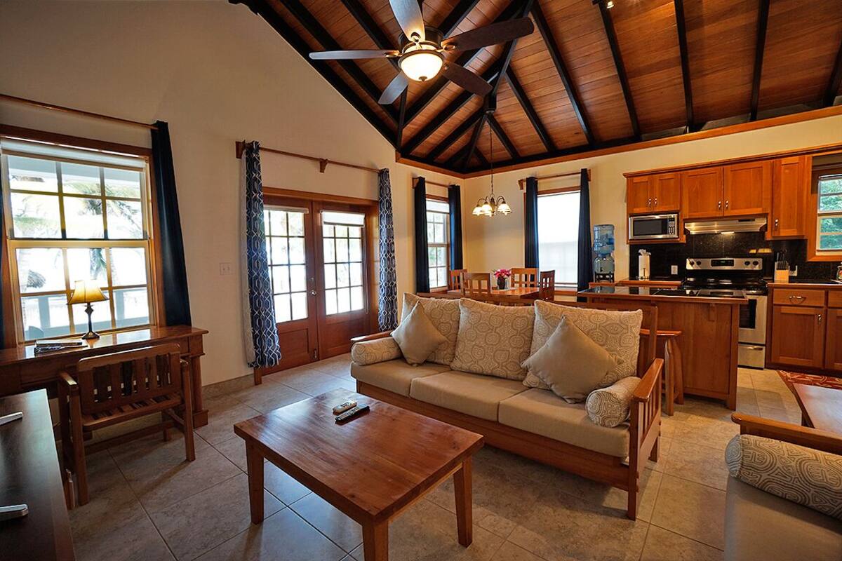 A spacious living area with a wooden ceiling, a sofa set, a coffee table, and a kitchen area with wooden cabinets and appliances.