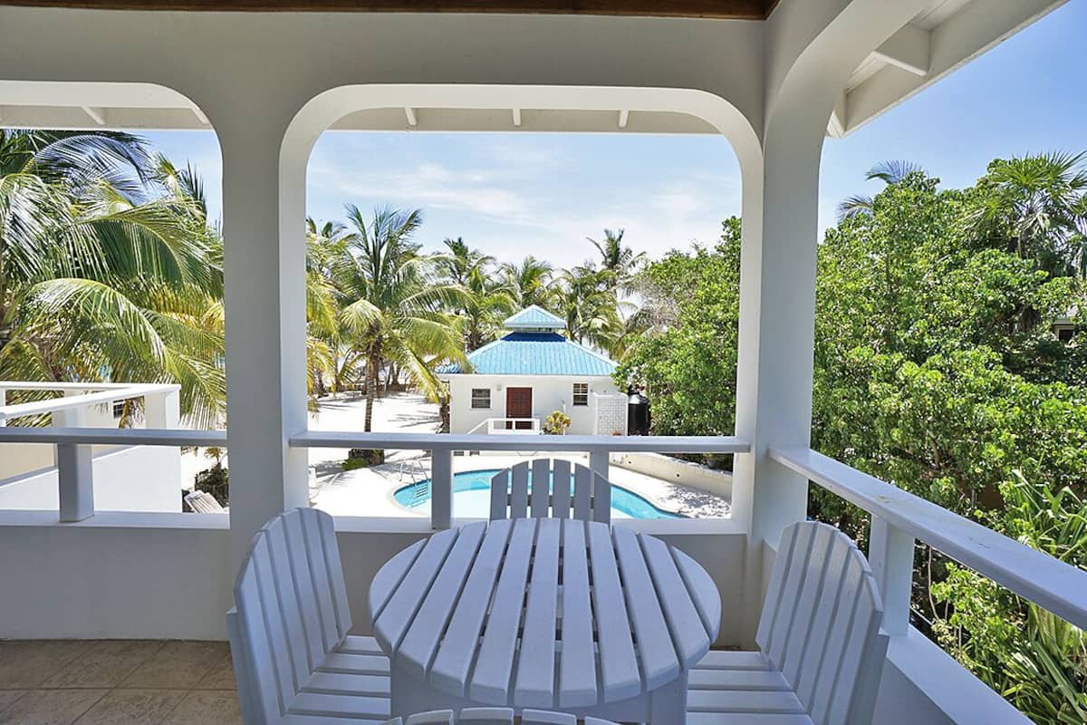 A balcony with a pool, white chairs, and a view of a blue-roofed building surrounded by palm trees.
