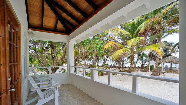 Un espace extérieur abrité, doté d’une balustrade blanche, d’une porte en bois et offrant une vue sur une plage bordée de palmiers.