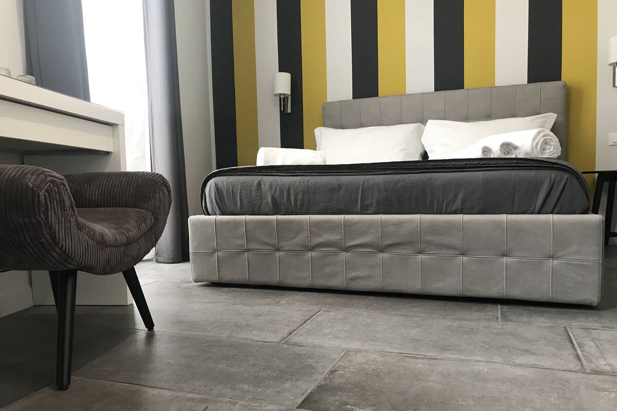 Un dormitorio moderno con una cama tapizada en gris, un sillón tapizado en gris y una pared a rayas en amarillo, negro y blanco.