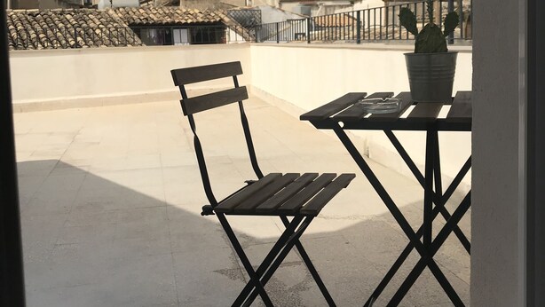 An outdoor seating area with a metal table and chair, a potted plant, and a view of historic buildings in the background. - B&B BaRock (Noto)