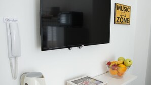 A white wall with a flat-screen TV, a white phone, a white kettle, a bowl of fruit, and a framed picture on a white shelf. - B&B BaRock (Noto)