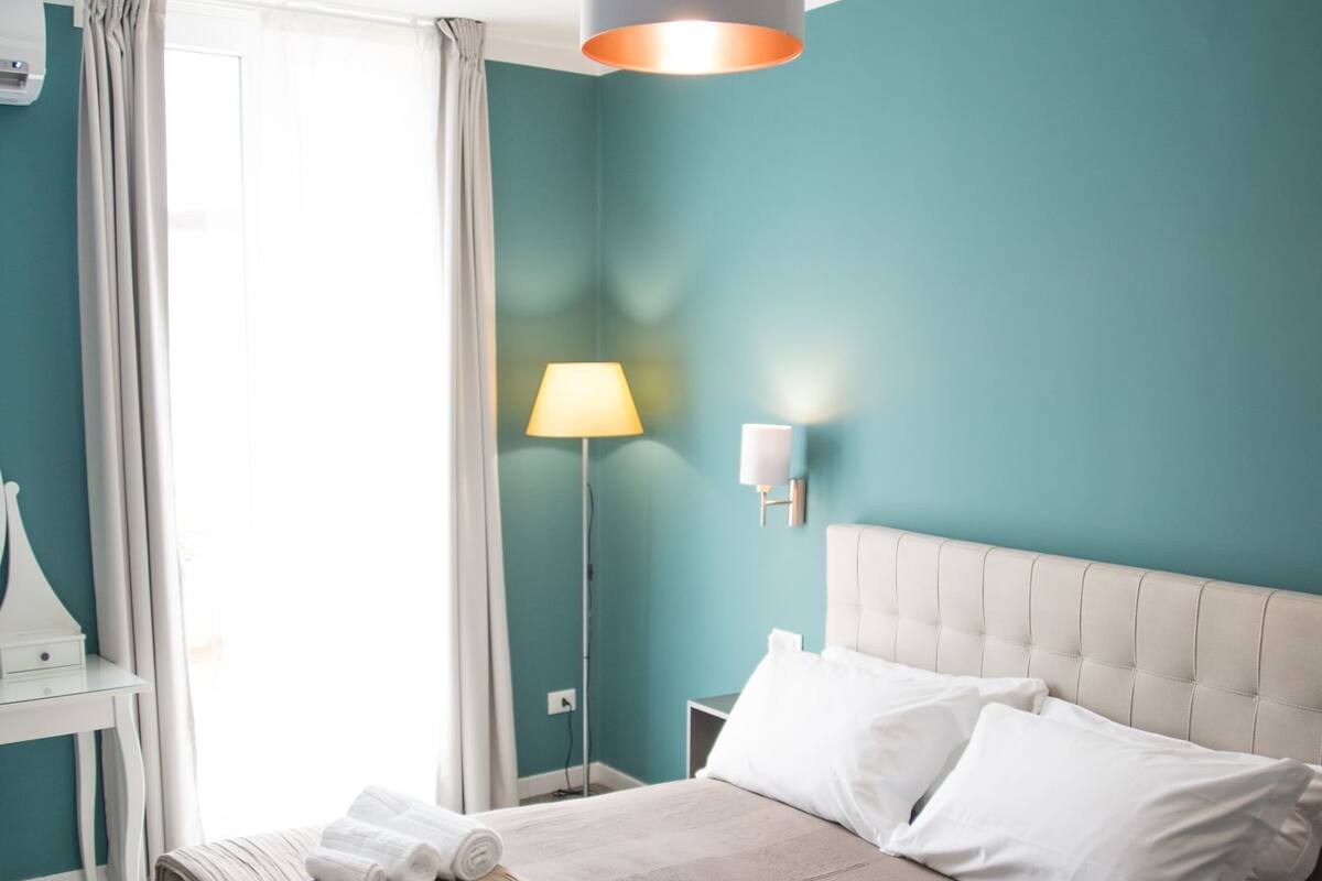Una cama bien hecha con sábanas blancas y una manta a rayas, una lámpara de noche y una lámpara de pared en una habitación con paredes color turquesa y techo blanco.