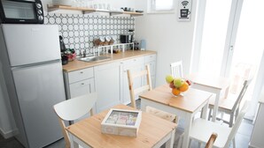 A modern kitchen with white cabinets, a microwave, and a sink. There are two wooden tables with chairs, one with a bowl of fruit. - B&B BaRock (Noto)
