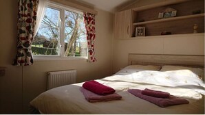 A bedroom with a large bed, a window with curtains, a radiator, and a wooden shelf with items. - Le Cabri Resort (Duras)