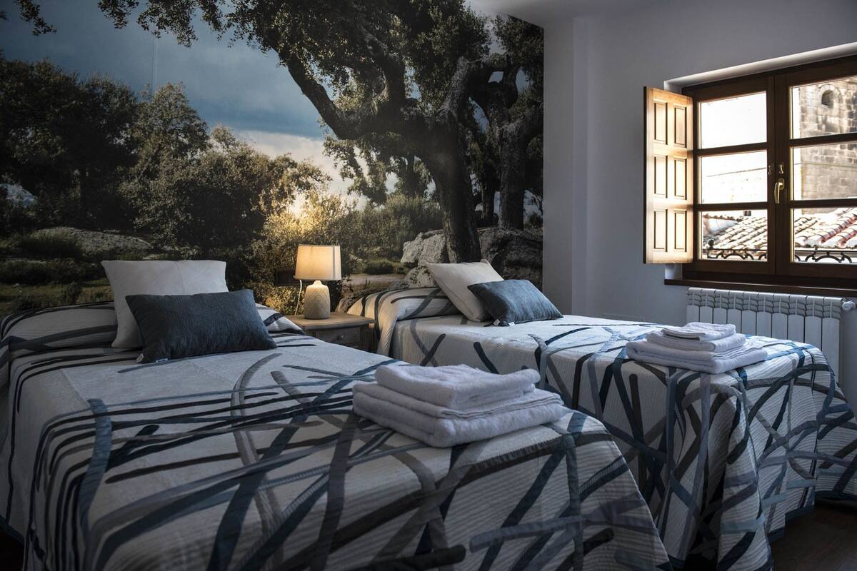 A bedroom with two beds, a mural of trees, a lamp, and a window with open shutters.