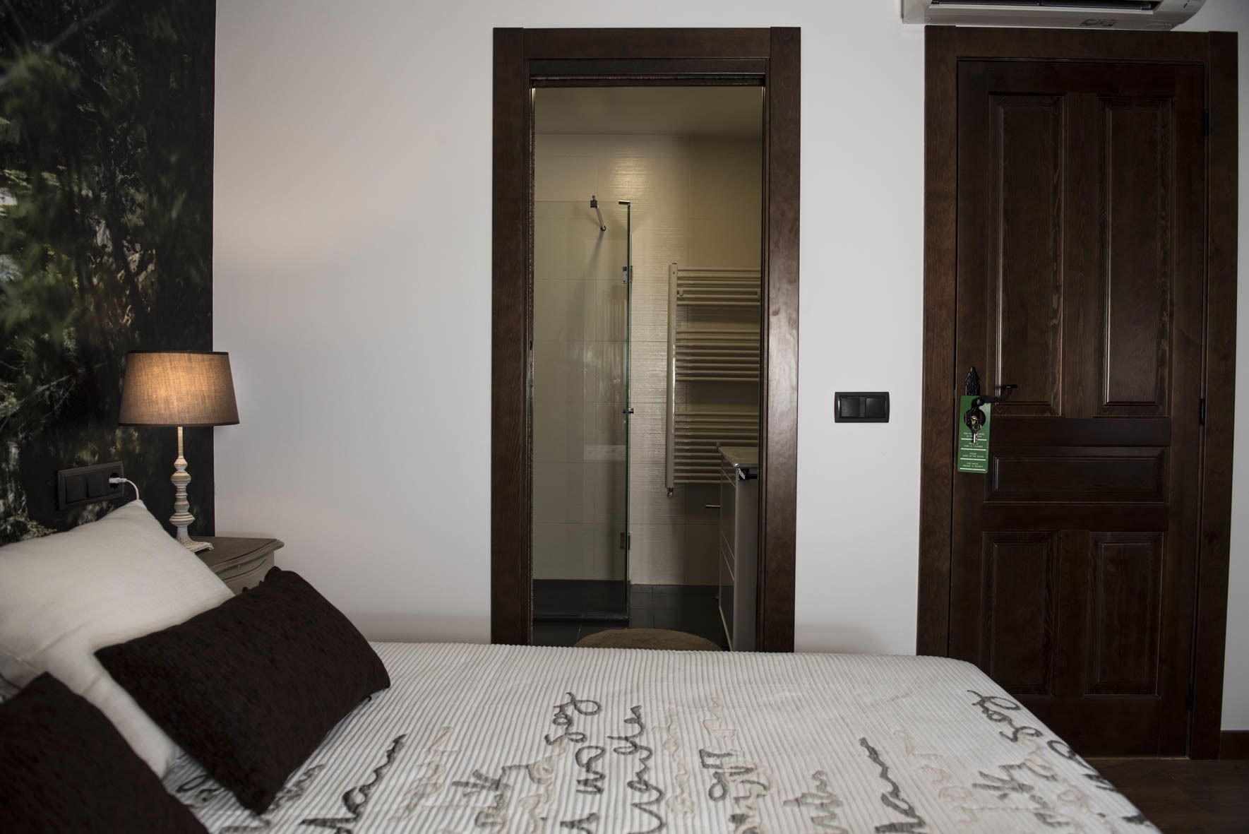 Una habitación de hotel con cama, mesita de noche con lámpara, puerta y vista al pasillo.