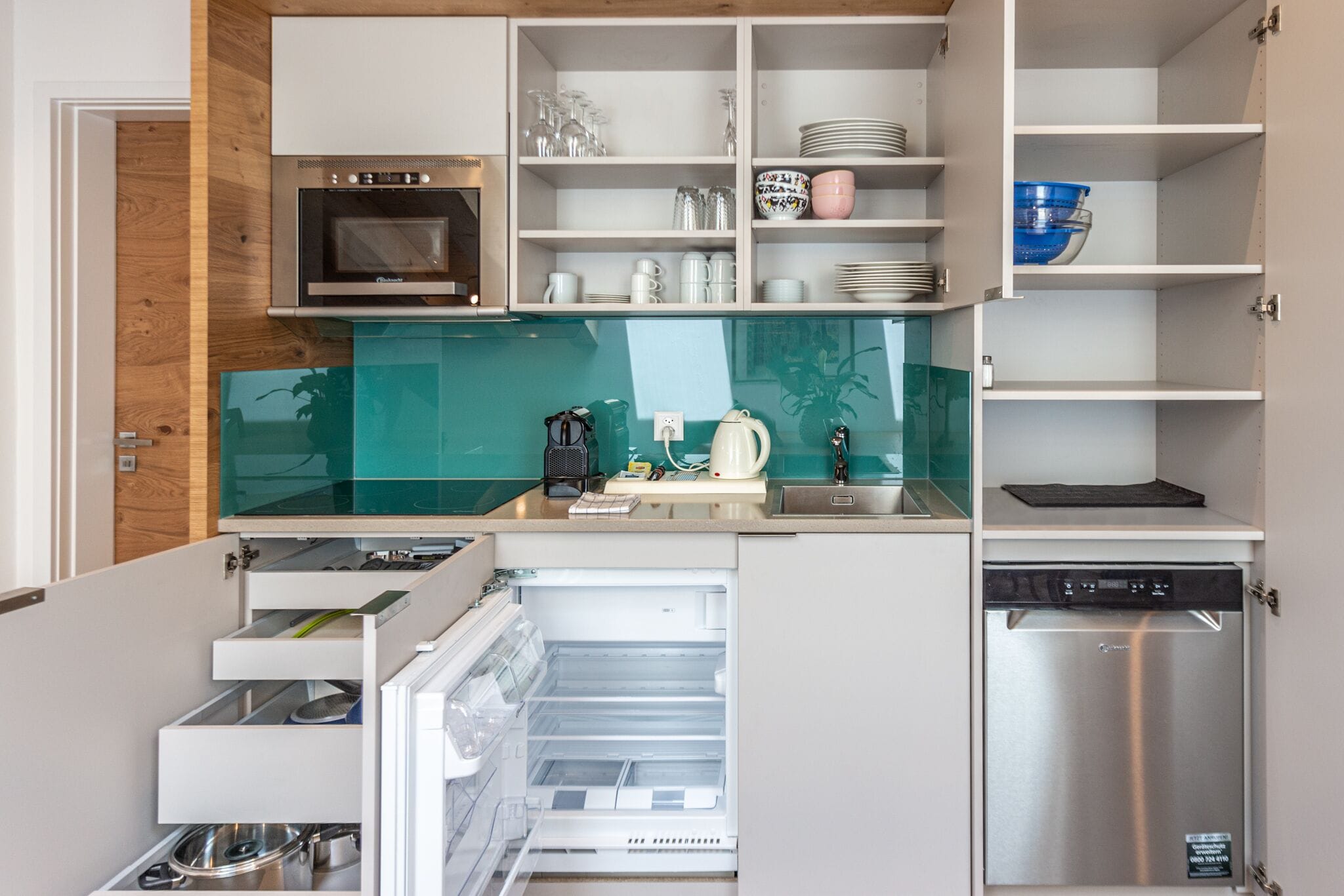 A compact kitchen with a built-in oven, microwave, and dishwasher. It includes open shelves with dishes and a small countertop area.
