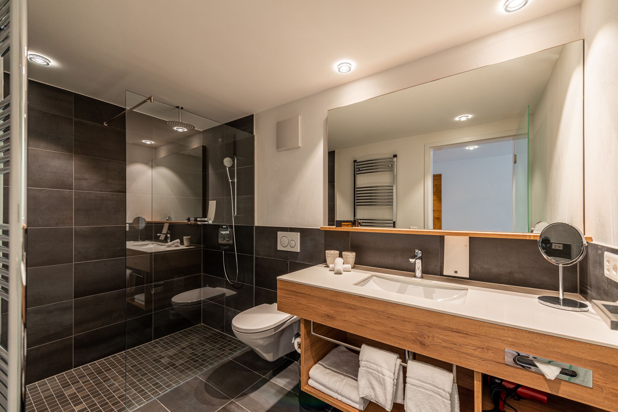 Een moderne badkamer met een douchecabine van glas, een wit toilet en een houten wastafelmeubel met een spiegel.