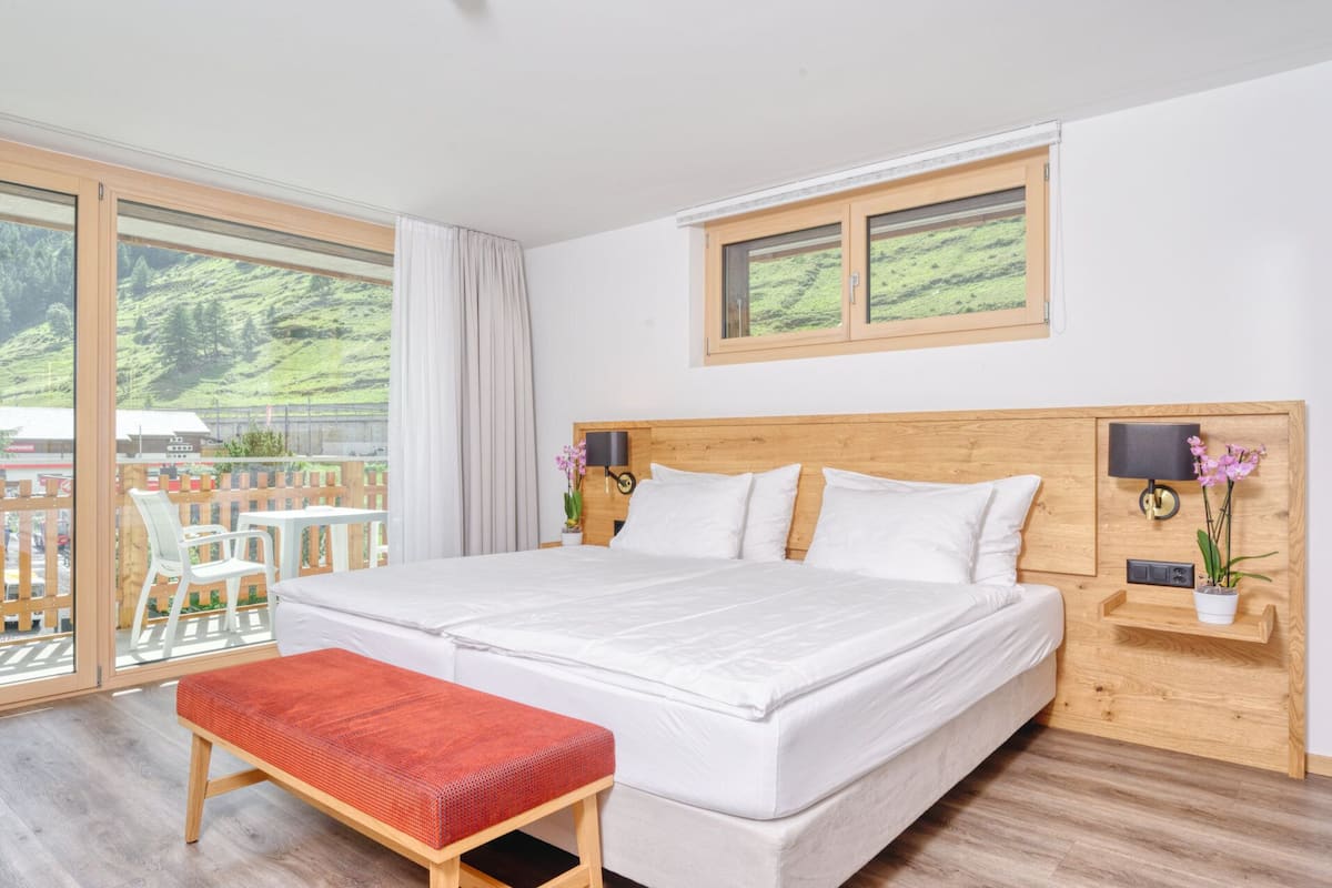 A large bed with a wooden headboard, a bench, and a view of a green landscape through large windows.