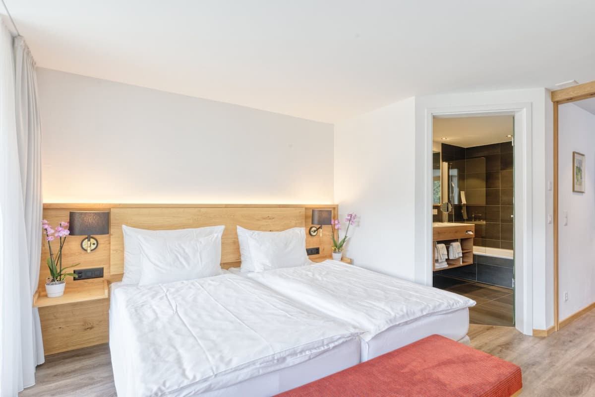 A modern hotel room with a large bed, wooden headboard, bedside tables, a red bench, and a bathroom with a dark tiled shower and a wooden vanity.
