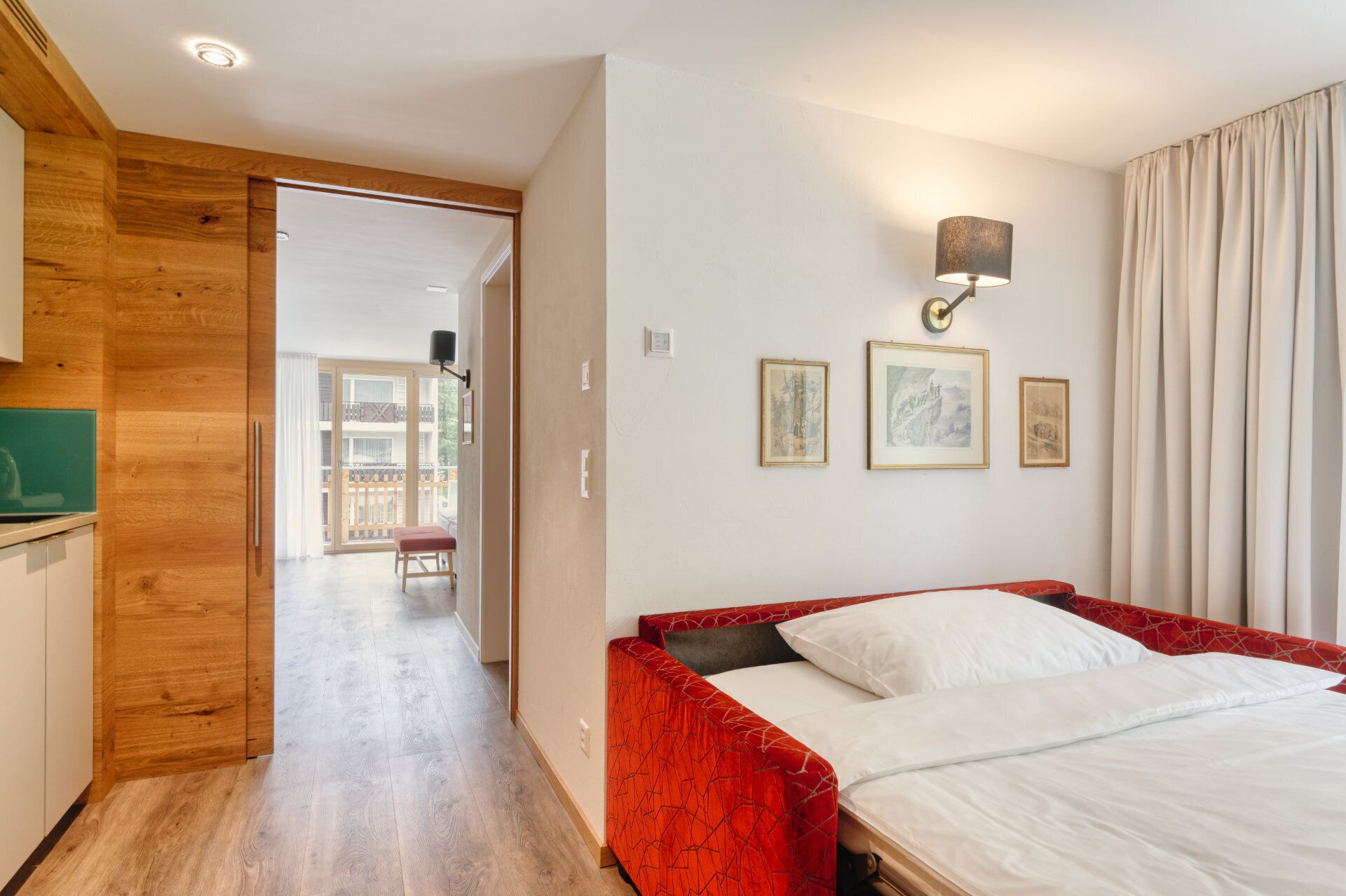 Une chambre d’hôtel avec un plancher en bois, un lit recouvert d’un tissu rouge, une lampe fixée au mur et des œuvres d’art encadrées.