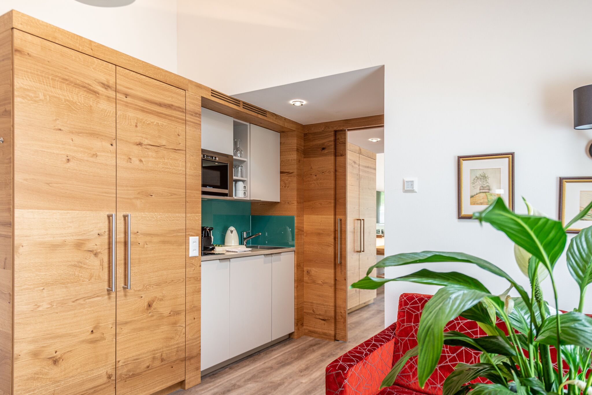 A modern kitchen with wooden cabinets, a microwave, and a sink. There is a red patterned sofa and a potted plant in the living area.