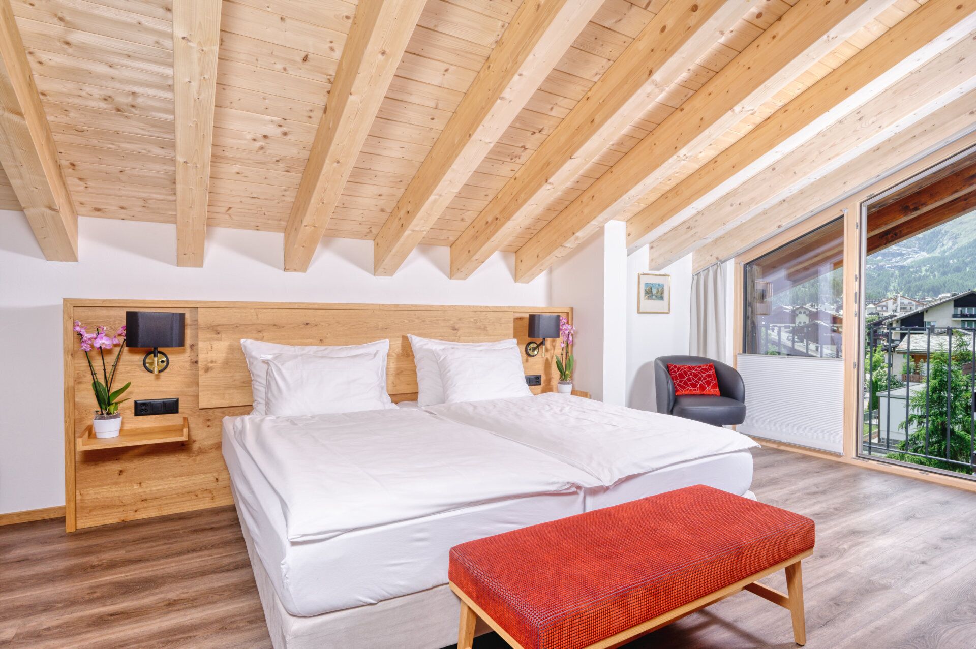 A bedroom with a wooden ceiling, a large bed, a bench, a chair, and a window with a view of a mountain.