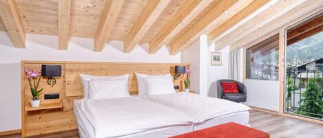 A bedroom with a wooden ceiling, a large bed, a bench, a chair, and a window with a view of a mountain.
