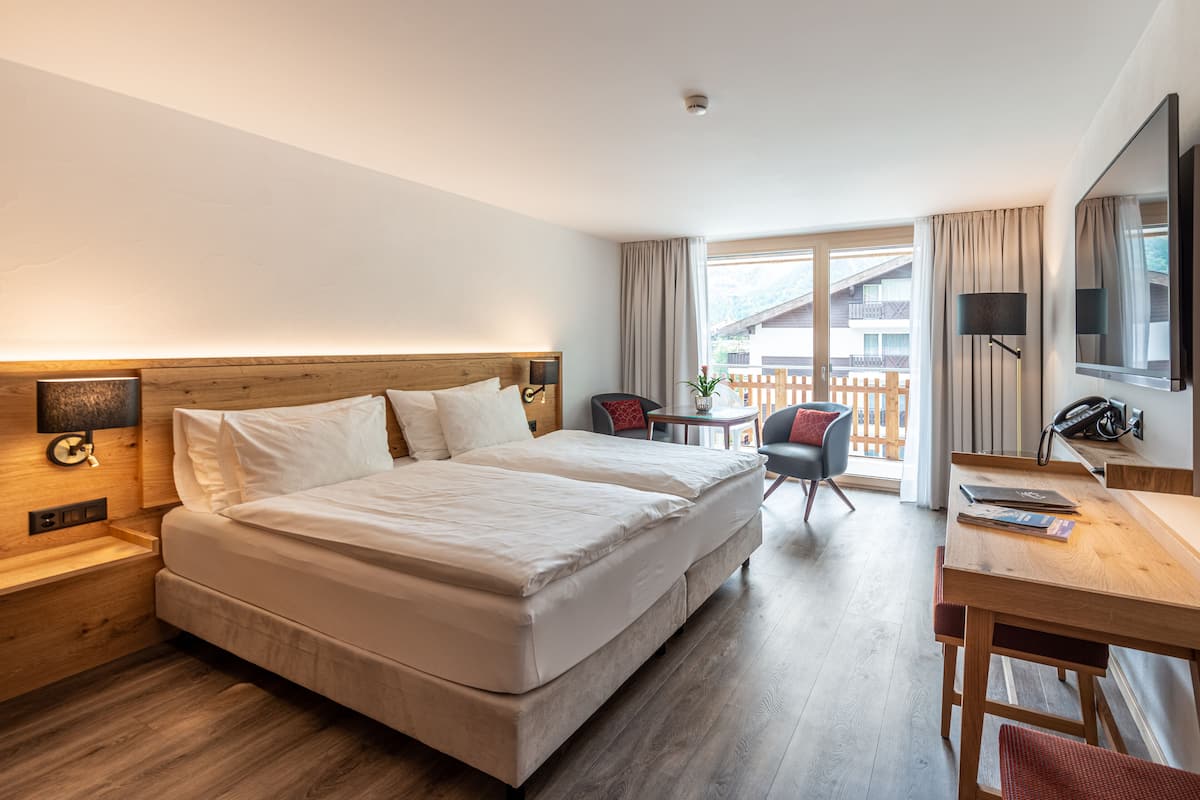 A hotel room with a double bed, a desk with a chair, a TV, and a balcony with a view of the mountains.