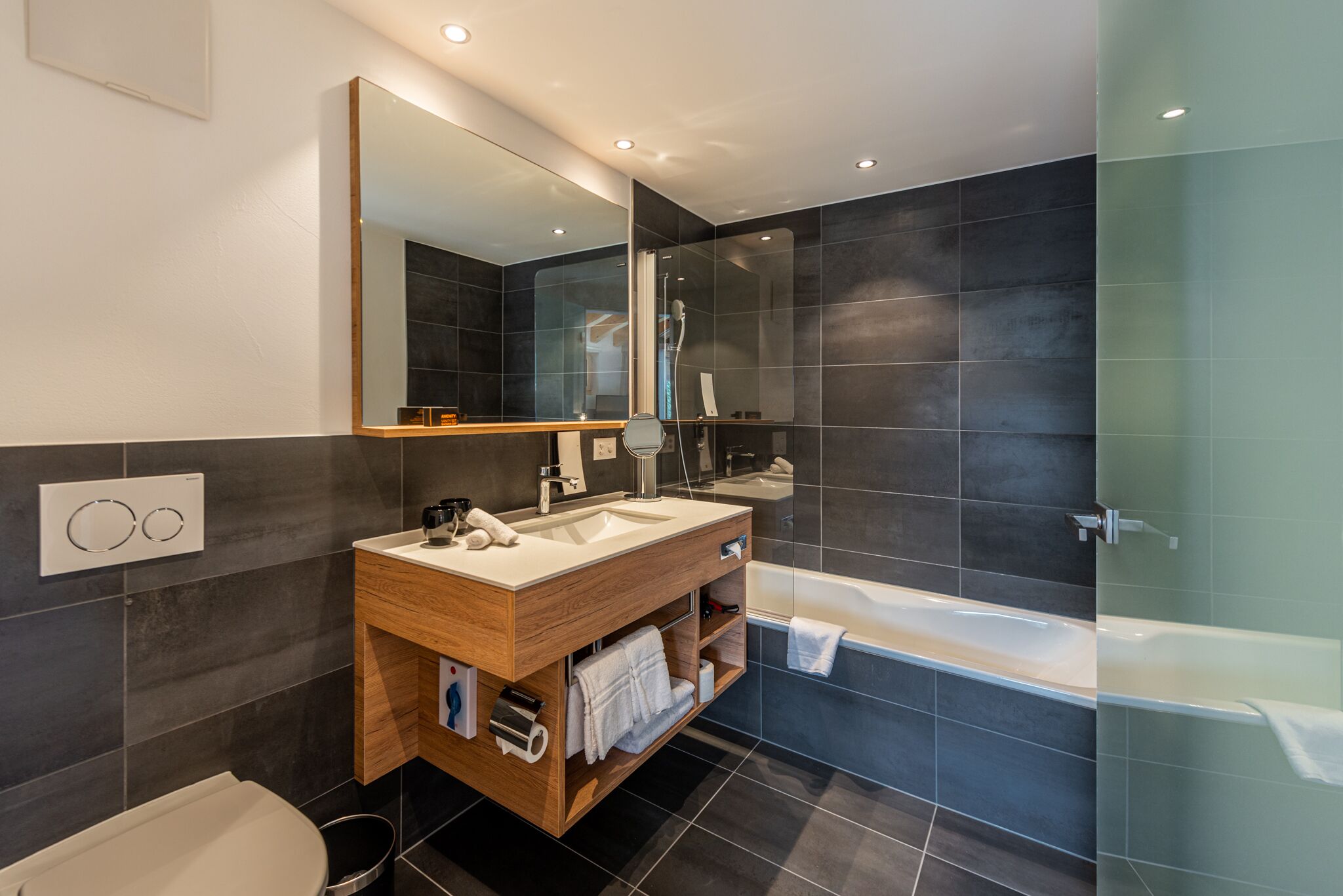 A modern bathroom with a wooden vanity, a large mirror, a bathtub, and dark tiled walls.