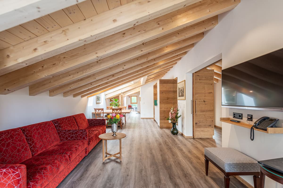 A modern loft-style living area with exposed wooden beams, a red patterned sofa, a small round table with a vase, and a bench.