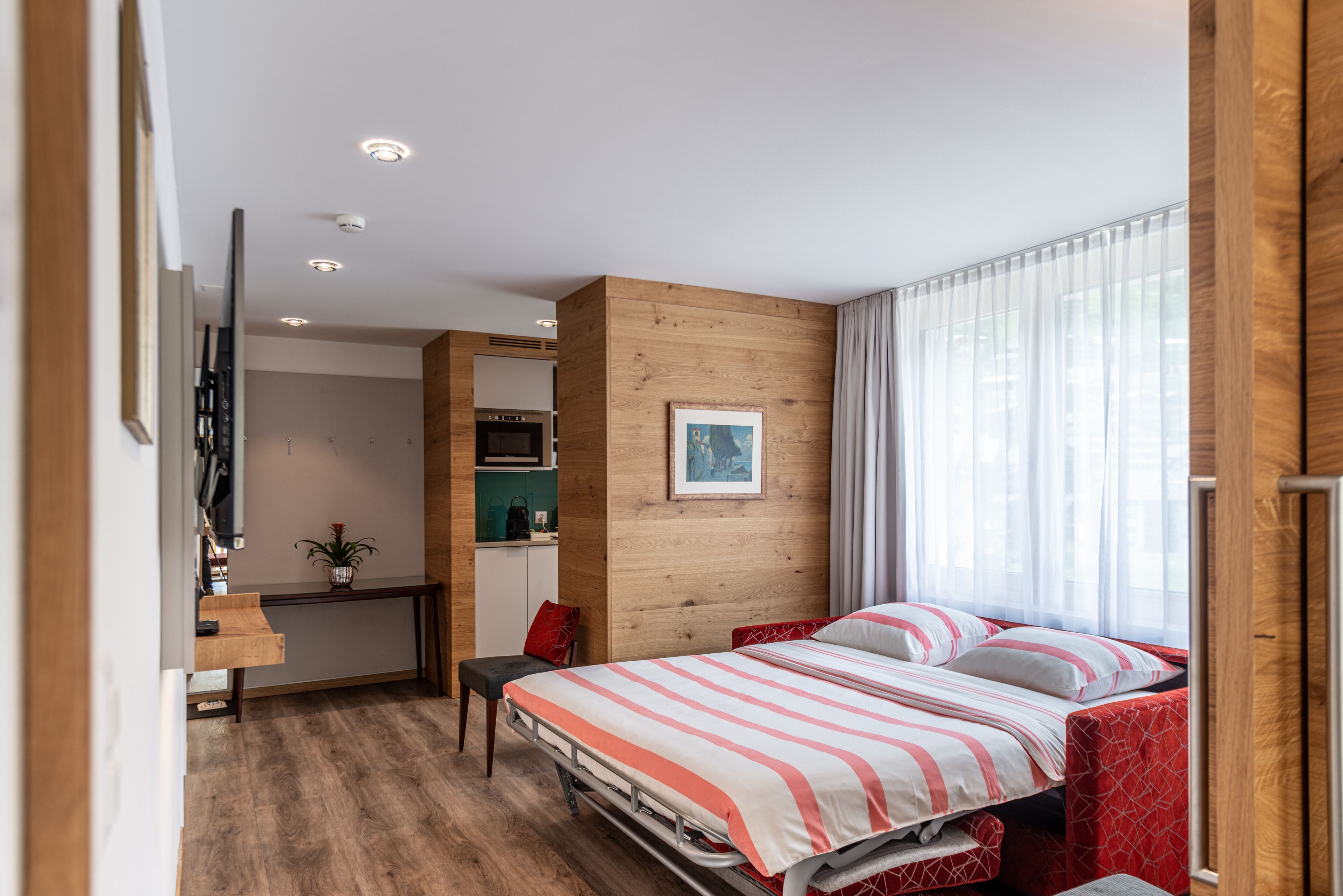 A modern hotel room with a wooden accent wall, a bed with striped bedding, a desk with a chair, and a view of the outdoors through a window.