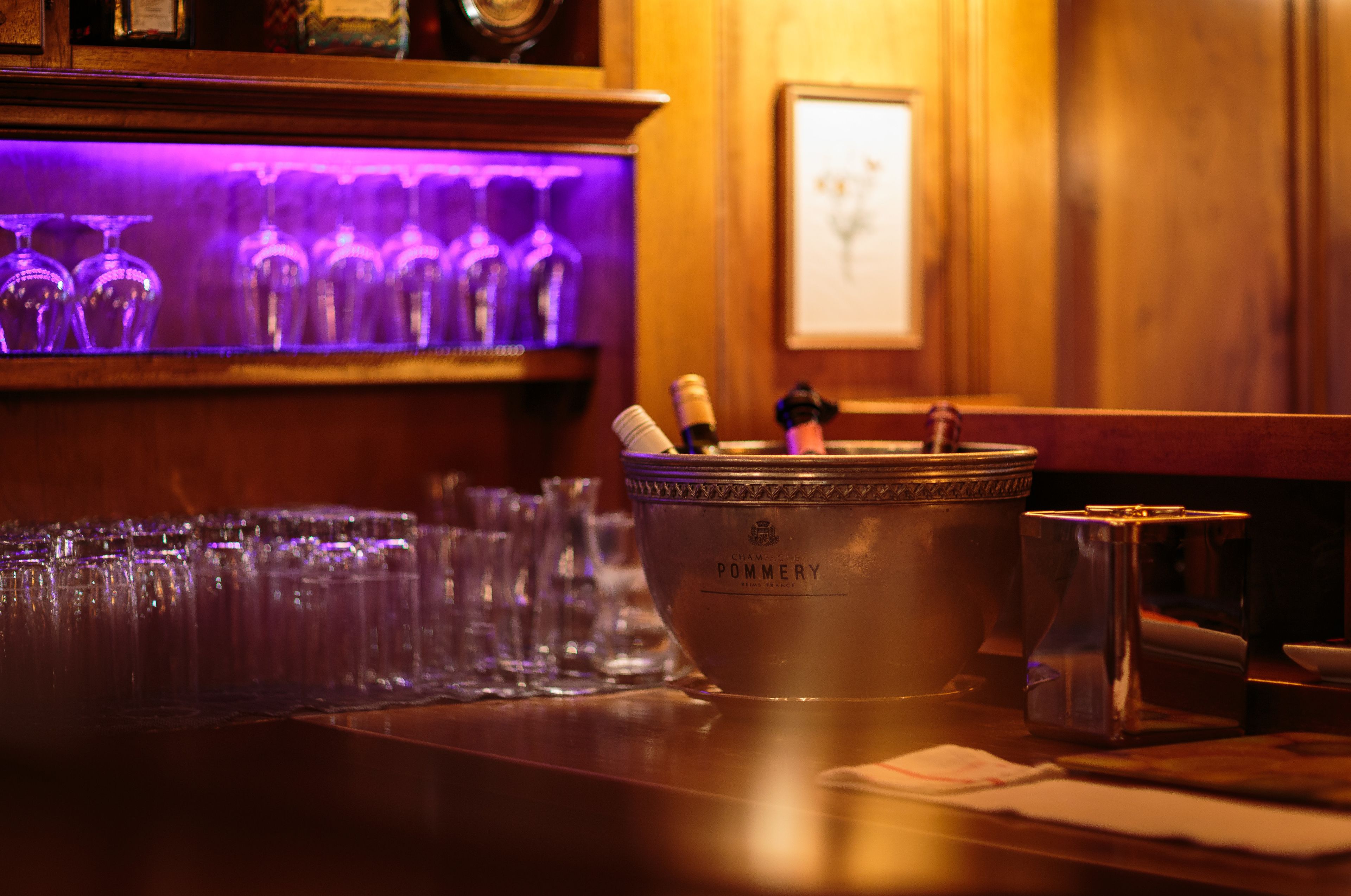 A bar area with a metal bucket containing bottles and glasses, a wooden bar counter, and a purple-lit shelf with multiple glasses.