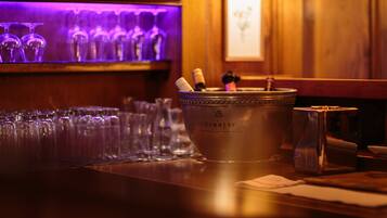 A bar area with a metal bucket containing bottles and glasses, a wooden bar counter, and a purple-lit shelf with multiple glasses.