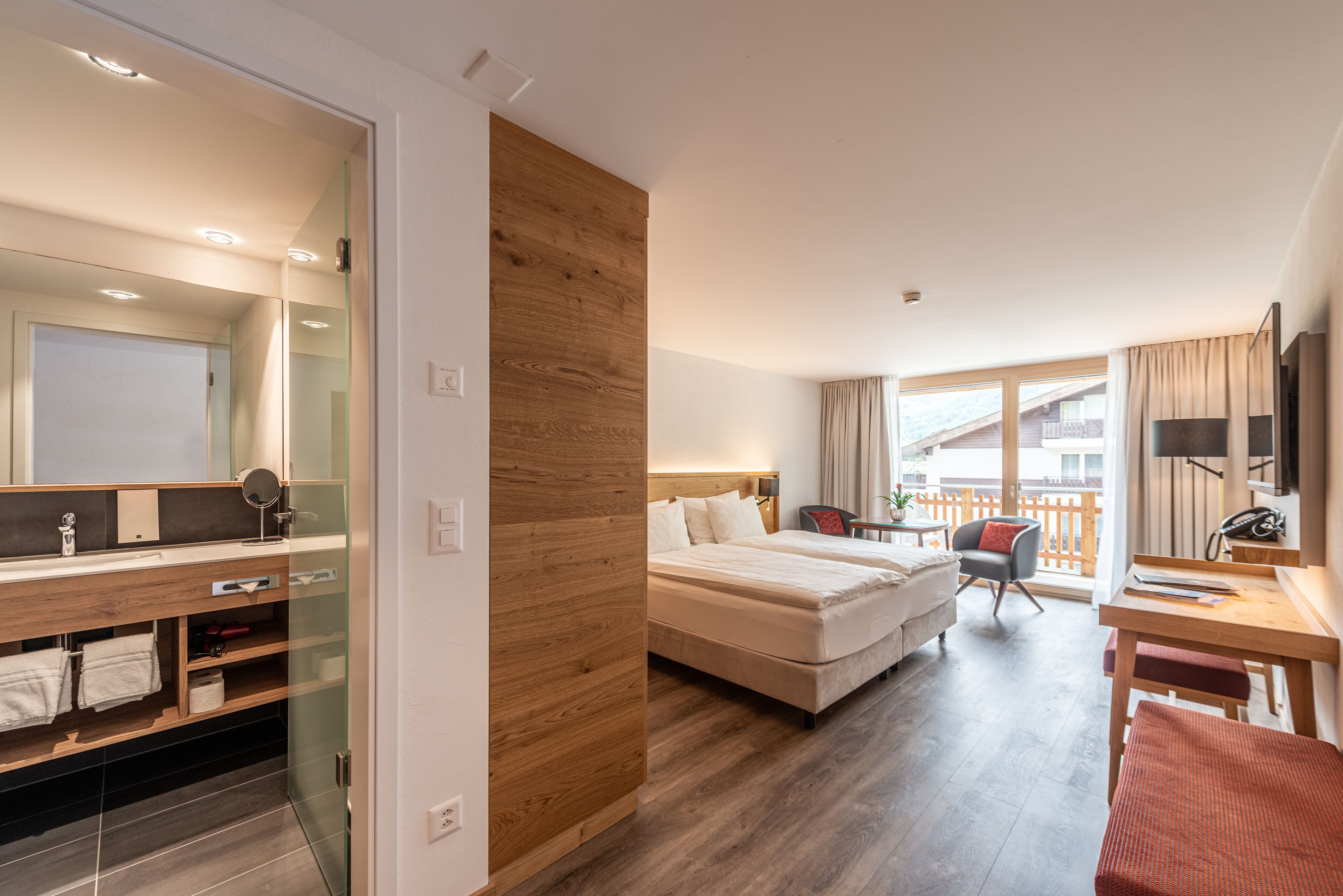 Ein Hotelzimmer mit einer Wandpaneinung aus Holz, einem großen Bett, einem Schreibtisch, einem Sessel, einem kleinen Balkon und einem Badezimmer mit Duschkabine.