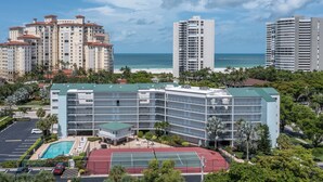 A multi-story hotel with a rooftop pool and tennis court, surrounded by palm trees and a beach view. - San Marco Residence 604- Across the Street from Beach, Hotels, Restaurants and Shopping!  Sleeps 6! (Marco Island)