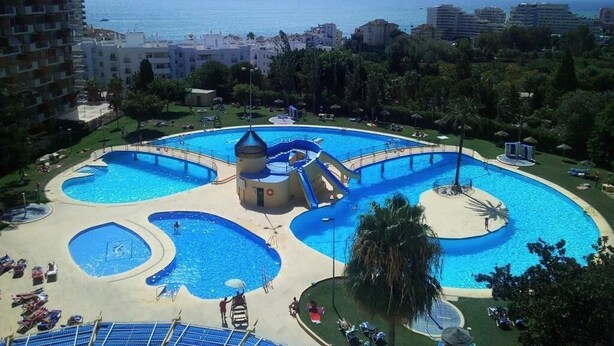 A large outdoor swimming pool with multiple sections and a water slide, surrounded by lounge chairs and palm trees, with a view of the sea and buildings in the background. - Cozy apartment with pool and sea views (Benalmádena)