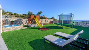 Een speelruimte voor kinderen met een glijbaan, schommels, een trampoline met veiligheidsnet, een zwembad en lounge fauteuils op kunstgras.