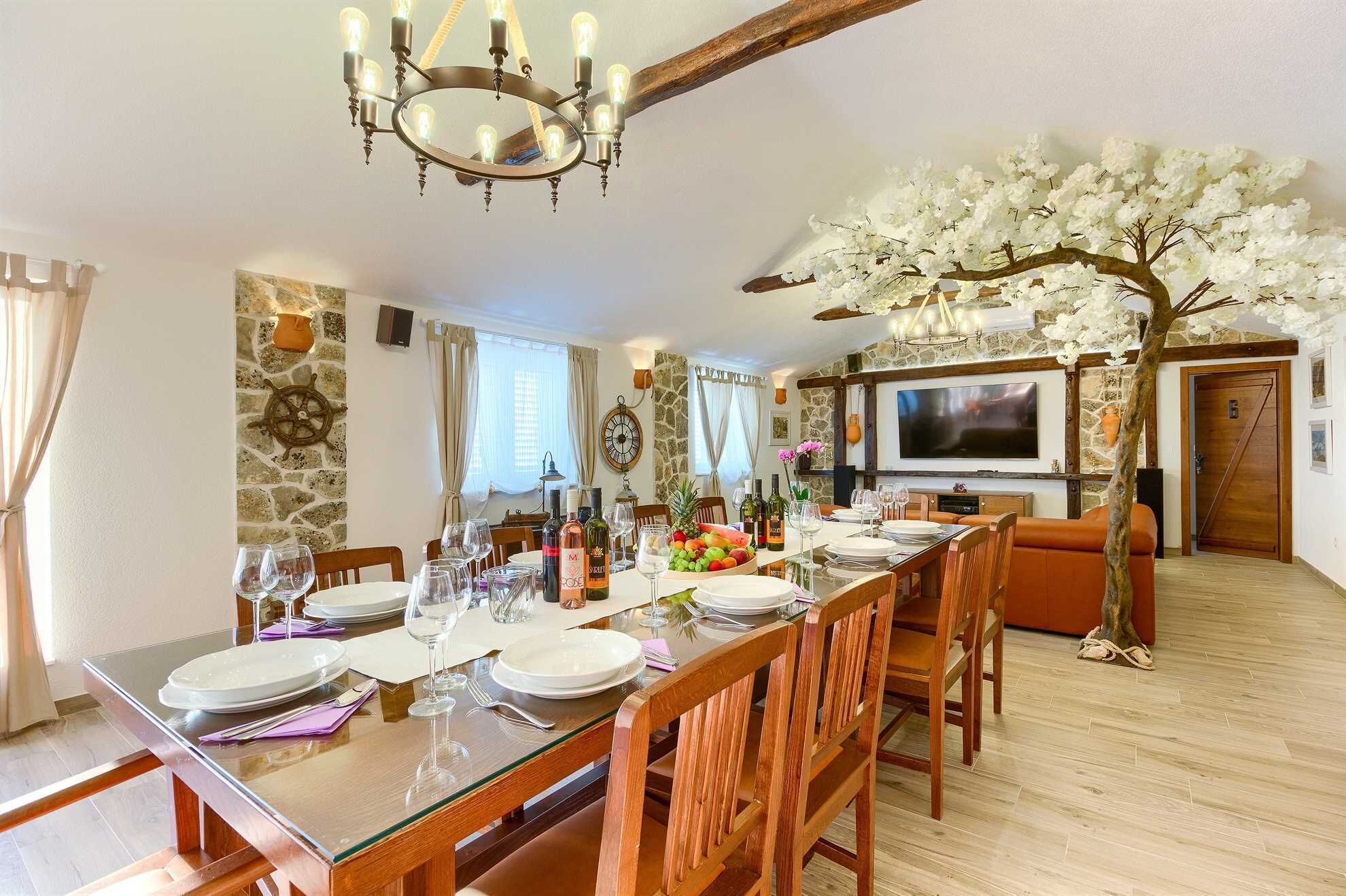 Ein Essbereich mit einem langen Holztisch, der für eine Mahlzeit gedeckt ist, einer Kristallleuchtre und einem Zierbaum mit weißen Blüten.