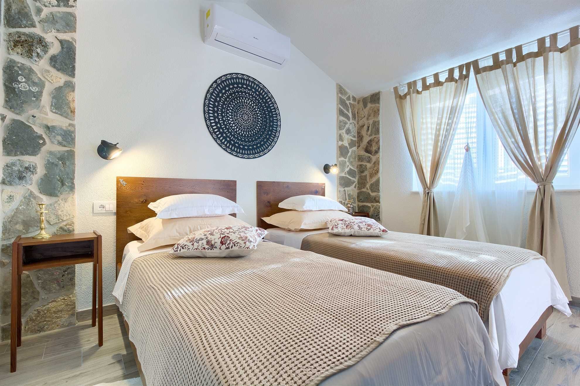 A room with two beds, stone walls, a decorative wall hanging, and a window with curtains.