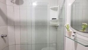 Une salle de bain moderne avec des carreaux blancs, une douche en verre, une étagère en verre où sont rangés les articles de toilette et un lavabo blanc.