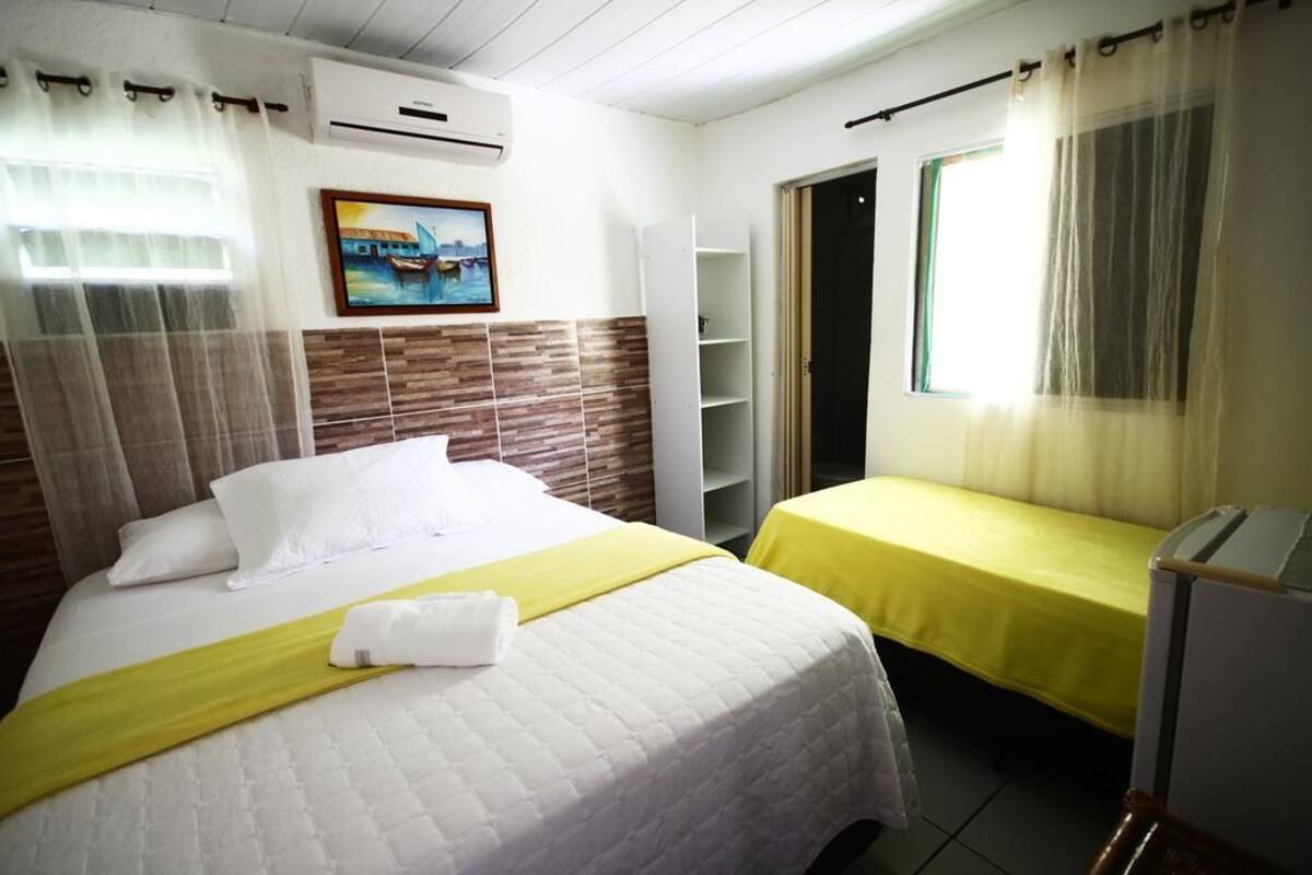 A hotel room with a bed, a bedside table, a window with curtains, a painting on the wall, and a door leading to another room.